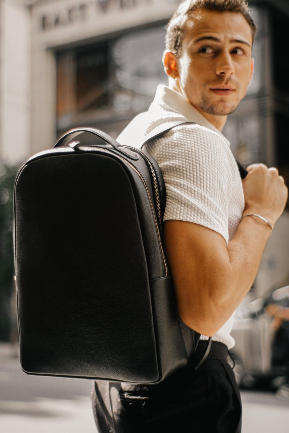 A man wearing a white short-sleeve shirt and black trousers carries a black leather backpack on his shoulder in an outdoor urban setting.